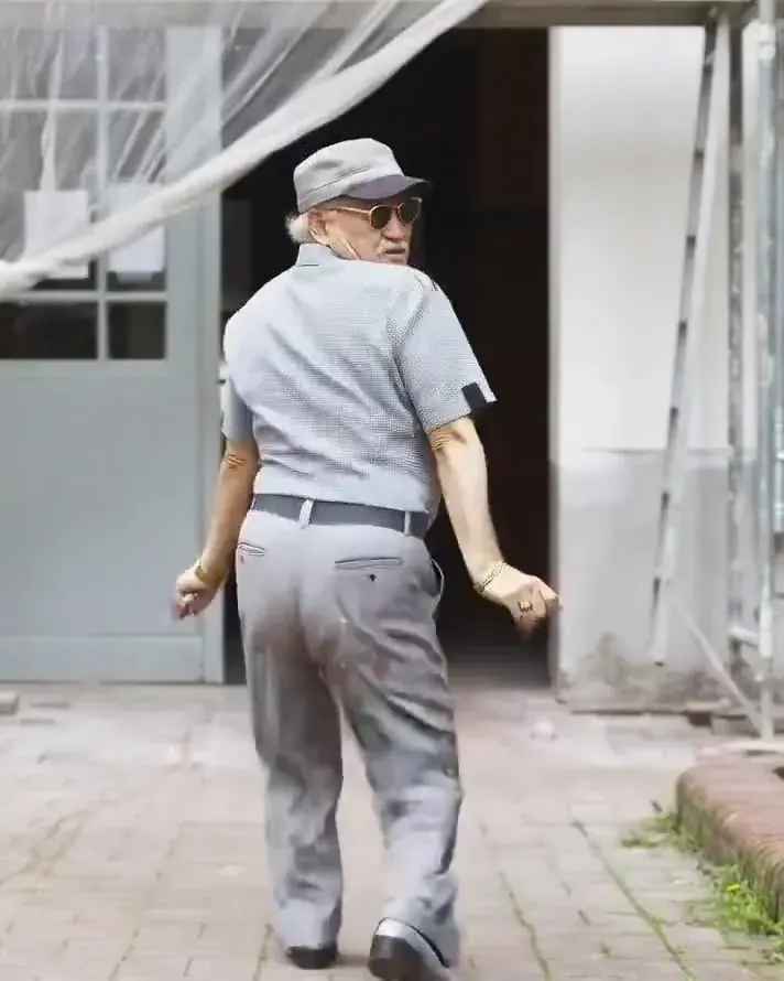 An elderly man in a cap, sunglasses and short-sleeved shirt dancing happily in a paved courtyard