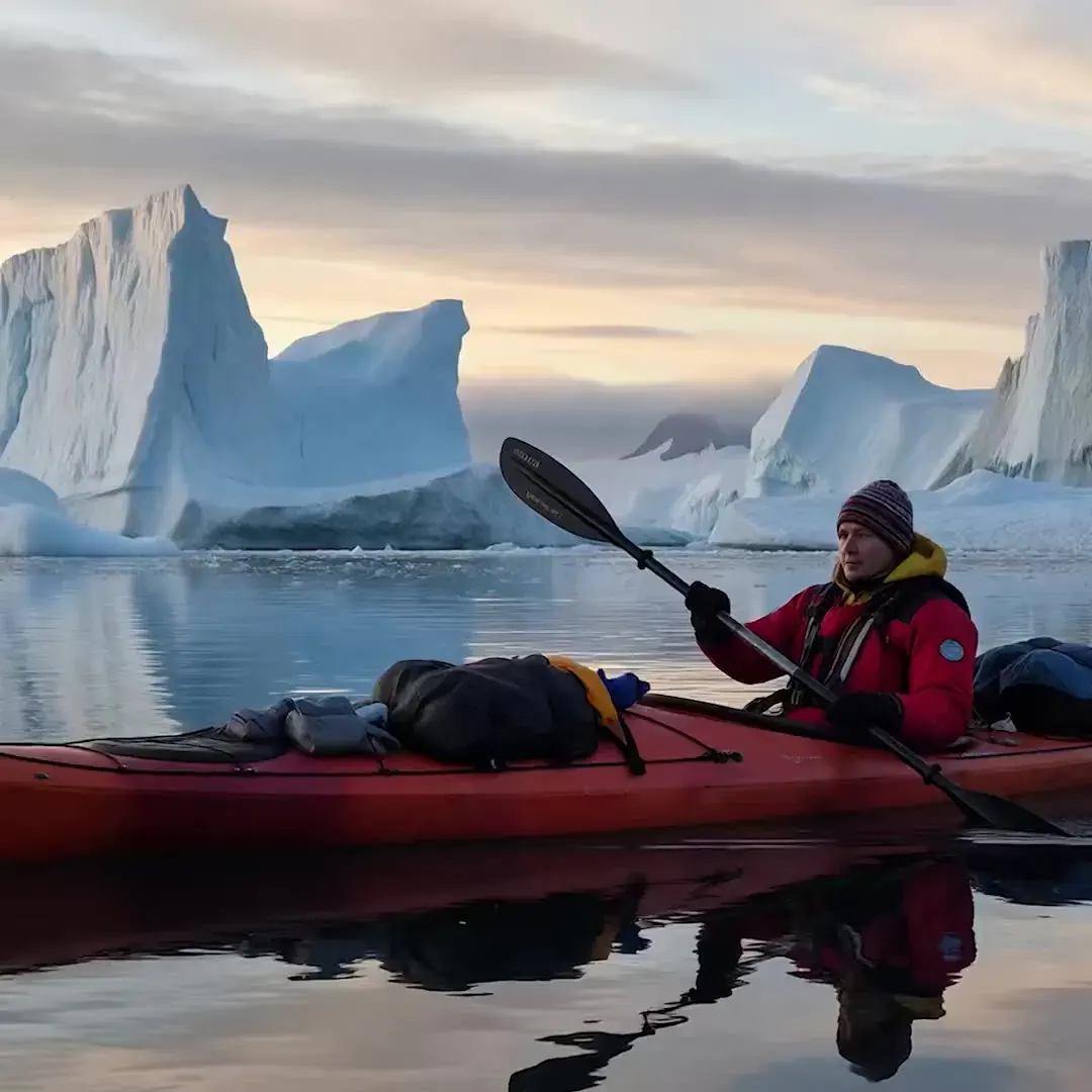 Veo 3 travel recipe illustration: a kayaker in a red jacket paddling between towering icebergs at dawn